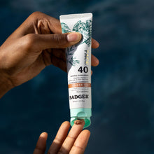 Load image into Gallery viewer, Hand holding a Badger Face Sunscreen tube against a blue background