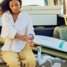Load image into Gallery viewer, Woman applying sunscreen to her arm with a surfboard and 50 SPF tube in the background.